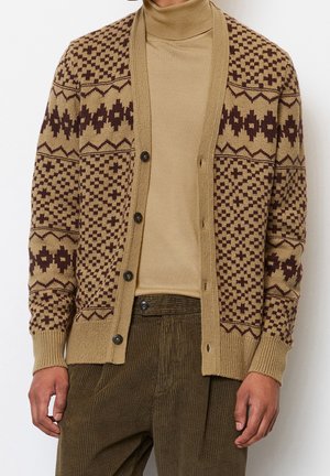 Mand iført en brun mønstret knap-op strikket cardigan over en beige rullekrave og olivengrønne kordfløjlsbukser.