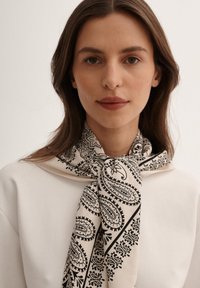 Kremowy szalik z czarnym wzorem paisley, starannie zawiązany wokół szyi. Gładka struktura, lekki materiał, z dekoracyjnym wykończeniem.