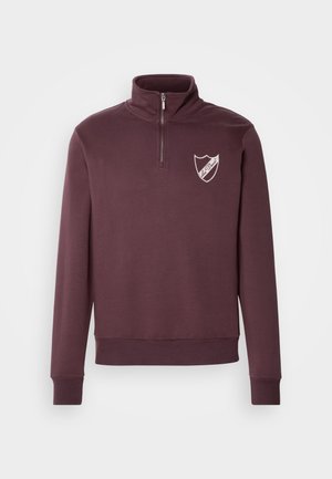 Bordo pulover s visokim ovratnikom, polu patentnim zatvaračem i bijelim grbom na lijevoj strani prsnog dijela. Mekana tkanina s rebrastim manžetama.