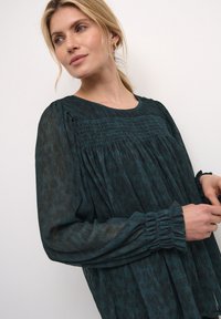 Blusa turchese con arricciature sul petto, maniche lunghe con dettagli arricciati e una vestibilità fluida realizzata in tessuto leggero e testurizzato.