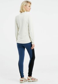 Ilga rankovė kreminė palaidinė, derinama su mėlynomis ir juodomis leggins. Modelis dėvi šviesius šlepetes; audinys lygus, o kirpimas prigludęs.