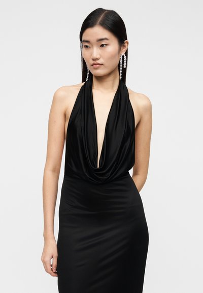 Nő hosszú sötét hajjal, fekete ujjatlan ruhában, mélyen kivágott, leomló nyakkivágással és hosszú, csillogó kristályfülbevalókkal.