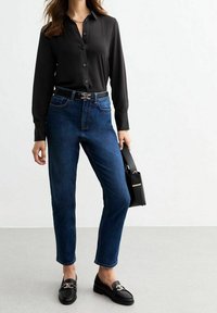 Chemise noire à manches longues, jean bleu foncé, mocassins noirs avec des accents dorés, et un sac à main noir avec un détail doré.