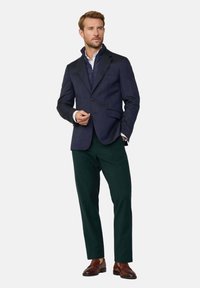 Blazer blu navy sopra un gilet grigio trapuntato, abbinato a pantaloni verde scuro. Scarpe eleganti marroni completano l'insieme. Camicia bianca visibile.