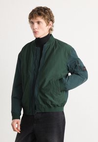 Jachetă bomber verde cu fermoar în față, guler ribetat și mansete elastice. Include o buzunar lateral și un patch cu logo pe mânecă.