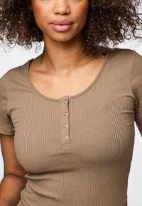 Približni prikaz žene odjevene u bež rebrasti top s kratkim rukavima i četiri srebrna drukera na prednjem izrezu oko vrata.