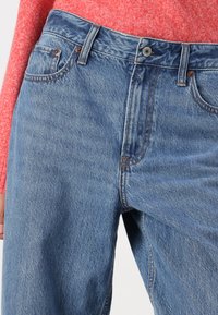 Un jean en denim bleu à taille haute, doté de quincaillerie en ton cuivre, de poches latérales et de légères déchirures pour un effet usé.