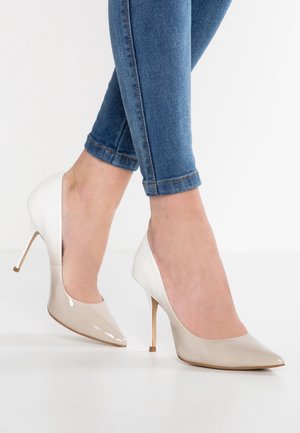 Kvinde iført beige laklæder højhælede sko med spidse tæer og blå skinny jeans mod en hvid baggrund.