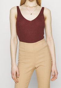 Vrouw in een bordeauxrode mouwloze gebreide top en high-waisted beige broek, geaccessoireerd met lagen gouden kettingen.