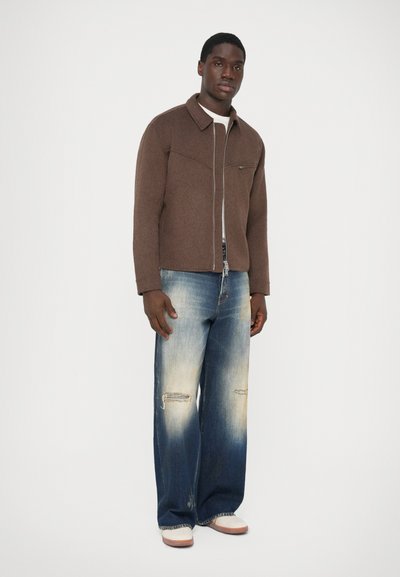 Uomo che indossa una giacca marrone con zip, una camicia bianca, jeans a gamba larga sbiaditi con strappi alle ginocchia e sneaker bianche su uno sfondo neutro.