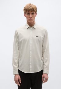 Ilgas rankovėmis dryžuotas marškinėlis šviesiai kreminės ir šviesiai rudos spalvų, su sagomis apykaklėje ir mažu juodu logotipu ant krūtinės. Lygios tekstūros.