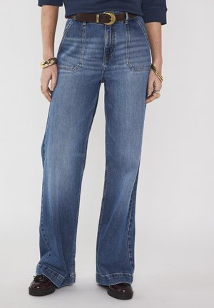 Jean bleu à jambes larges avec grandes poches à l'avant, associé à une ceinture foncée, des chaussures noires épaisses et des bijoux en or aux poignets et aux doigts.
