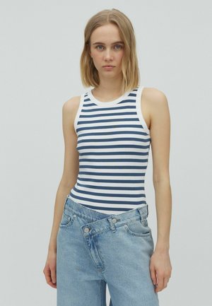 Femme aux cheveux blonds mi-longs portant un haut sans manches rayé bleu marine et blanc et un jean bleu clair déboutonné sur un fond uni.