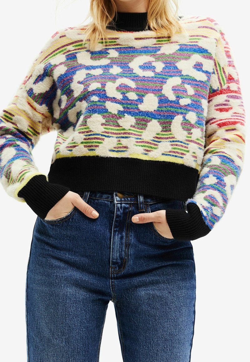Femme portant un pull en peluche à motifs colorés avec des poignets et un ourlet côtelés noirs, associé à un jean bleu taille haute, mains dans les poches.