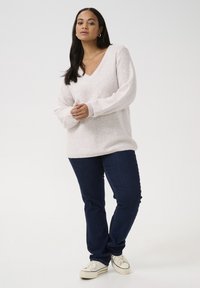 Pulover roz deschis, cu decolteu în V, cu un finisaj texturat și accente fine de sclipici, asortat cu blugi bleu închis și adidași albi.