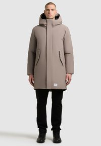 Cappotto imbottito beige leggero con cappuccio, colletto alto, due tasche laterali e una patch con logo all'orlo, abbinato a pantaloni neri e stivali.