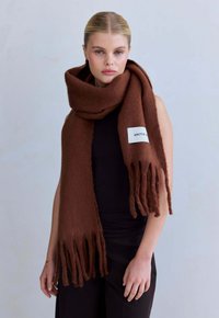 Bruine wollen sjaal met franjes, in rechthoekige vorm en met een label van stof met de tekst "ARCTIC", gedragen om de nek.