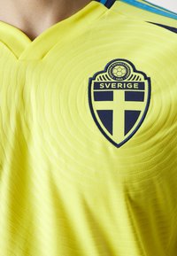 Żółta koszulka piłkarska z prążkowaną fakturą, z niebiesko-żółtym herbem z napisem "SVERIGE" na środku. Zapinana na dekolt w serek.