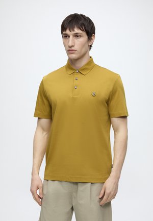 Mężczyzna ubrany w polówkę o krótkim rękawie w kolorze musztardowym z beżowymi spodniami, stojący na jasnym, jednolitym tle.