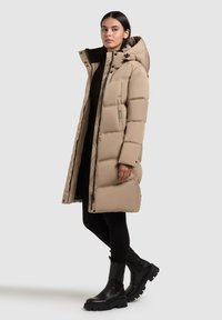 Jauna sieviete valkā garu bēšu izšūtu lietusmēteli ar kapuci, melnas legingi un melnas masīvas zābakus, stāvot sāniski uz vienkārša fona.