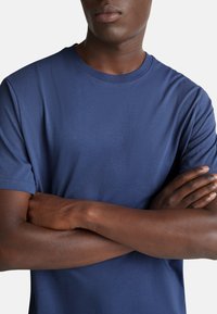 Námořnická modrá tričko s krátkým rukávem vyrobené z měkkého, hladkého materiálu; střih s kulatým výstřihem a přiléhavým siluetou. Model s překříženýma rukama.