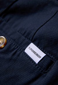 Marineblauwe stof met een gladde textuur, voorzien van een grote gouden knoop en een wit label met de tekst "LINDBERGH" in het zwart.