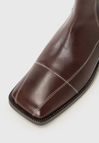 Botte en cuir marron avec un embout carré, ornée de coutures blanches et d'une semelle noire robuste, positionnée sur fond blanc.
