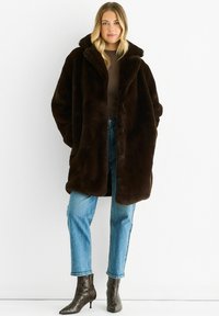 Brown faux fur coat with a large collar, button closure, and oversized fit, paired with light blue jeans and patterned ankle boots.