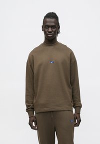 Felpa marrone in tessuto strutturato, con collo rotondo, spalle scese e una piccola targhetta con logo blu sul petto.