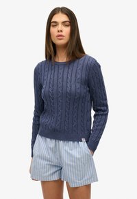 Navy blauwe kabel gebreide trui met een ronde halslijn en geribde manchetten, gecombineerd met lichtblauwe streepjesshorts. Opvallende textuur in het breiwerk.