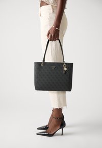 Sac en cuir texturé noir au design structuré, avec des accents en métal doré et un pendentif. Associé à un pantalon crème et des talons noirs.