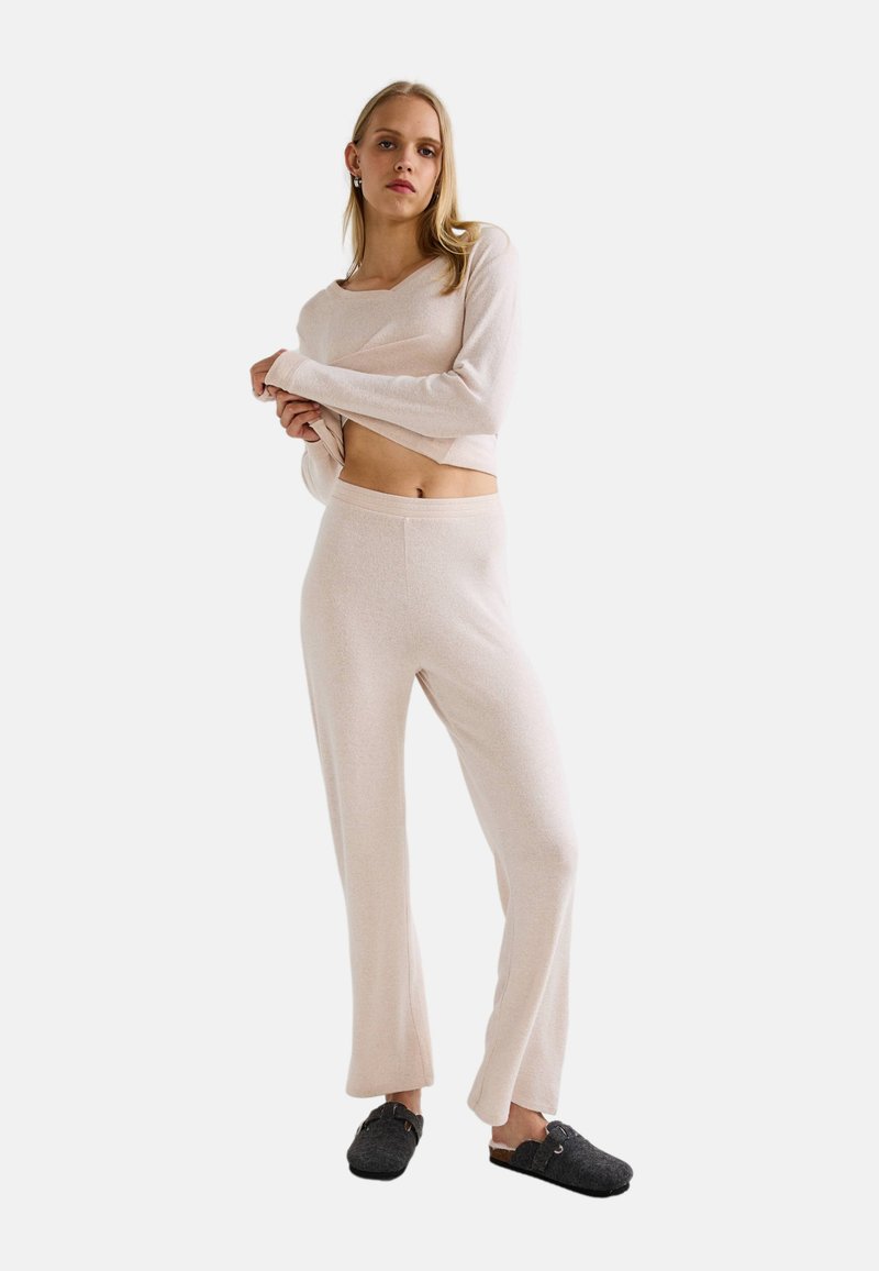 Femme portant un haut court à manches longues beige et un pantalon assorti, soulevant légèrement son haut, se tenant sur un fond blanc avec des chaussures noires à enfiler.