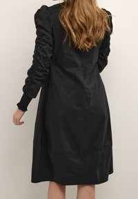 Vestido negro con mangas abullonadas y textura fruncida en los hombros. Longitud por debajo de la rodilla, tejido suave, sutil detalle de costura en la parte trasera.