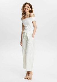 Haut blanc à épaules nues en maille rayée associé à un pantalon large taille haute crème avec une ceinture en tissu et des poches discrètes. Sandales à talons.
