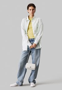 Camicia bianca oversize, canotta gialla a costine, jeans azzurri larghi, sneakers bianche e una piccola borsa bianca con tracolla a catena.