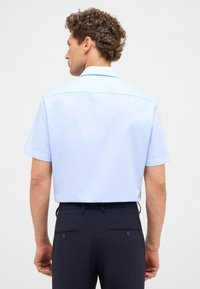 Camicia azzurro chiaro a maniche corte con colletto, texture liscia e vestibilità rilassata, abbinata a pantaloni scuri con doppie tasche posteriori.