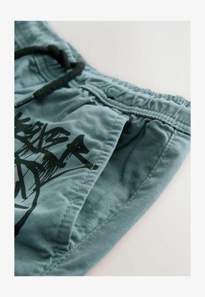 Vaaleansiniset lyhyet housut, jotka on valmistettu pehmeästä kankaasta. Niissä on musta graafinen printti edessä ja joustava vyötärönauha, jossa on kiristysnauha.