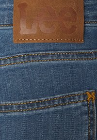 Tejido de mezclilla azul con costuras amarillas de contraste y una etiqueta de cuero marrón con el mensaje "Lee" en relieve. Los detalles incluyen costuras en los bolsillos y textura.