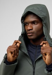 Dark green jacket with a drawstring hood, two-way zipper, and elastic cuffs. Worn over a navy mock-neck top, showcasing a matte texture.