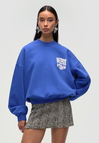 Sudadera oversized azul con texto gráfico blanco, cuello redondo acanalado y puños elásticos, combinada con una falda mini de estampado.