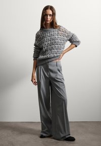 Maglione grigio lavorato a maglia con un motivo a rombi, abbinato a pantaloni grigi a gamba larga con una piega. Scarpe nere completano l'outfit.