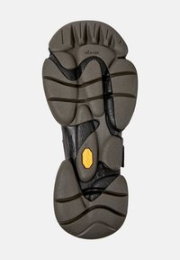 Sula av en Vibram-sko med texturerade, segmenterade greppmönster i svart och grått, med en gul Vibram-logga nära mitten.
