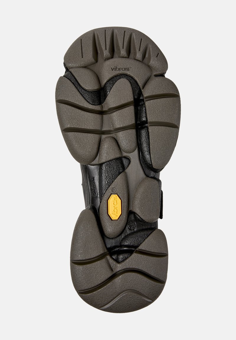 Semelle d'une chaussure Vibram avec des motifs de grip texturés et segmentés en noir et gris, comportant un logo Vibram jaune près du centre.