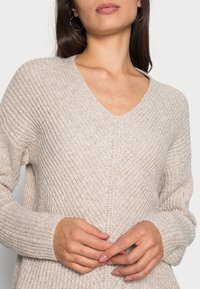 Pull en maille beige avec col en V, poignets côtelés et motif texturé. Comprend des manches longues et une coupe décontractée, mettant en valeur un tissu doux et chaud.
