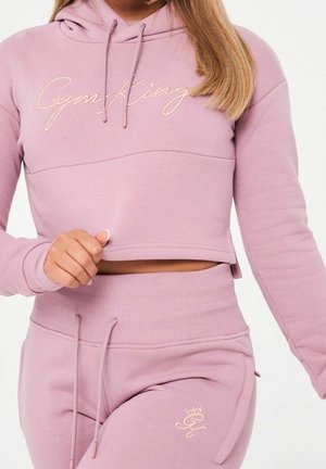 Femme portant un hoodie court rose poussiéreux assorti et un pantalon de jogging taille haute avec cordons de serrage et logo brodé "Gym King".