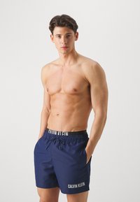 Șorturi de baie bleumarin cu talie elastică, șnur și logo-ul "CALVIN KLEIN" în alb. Textură netedă, design până la genunchi.