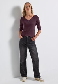 Bordeaux rood langemouwen bovenstuk met een V-hals en knopen, gedragen met losse zwarte jeans. Model staat met de handen in de zakken tegen een witte achtergrond.