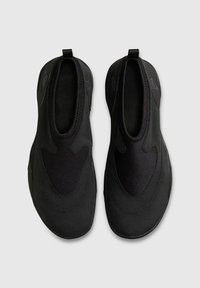 Ζευγάρι μαύρα αθλητικά παπούτσια τύπου slip-on με υφασμάτινη υφή, στρογγυλεμένες μύτες και θηλιές στα πίσω μέρη, σε θέα από πάνω σε γκρι φόντο.