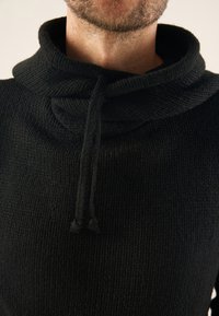 Sweat à capuche noir en maille avec un col montant et des cordons de serrage. Tissu texturé visible, mettant en valeur des motifs de point complexes et une coupe ajustée.