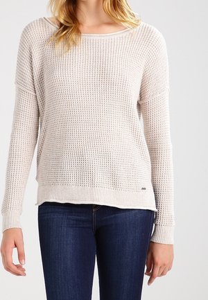 Woman wearing a light beige waffle-knit sweater with long sleeves and dark blue jeans, standing against a plain background.